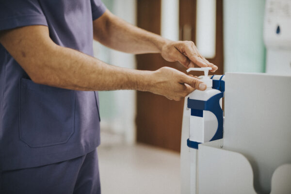 "Profesional sanitario con uniforme morado utilizando un dispensador de gel desinfectante en un entorno clínico."