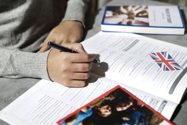 Persona escribiendo en un cuaderno de inglés, con apuntes y un bolígrafo sobre la mesa.